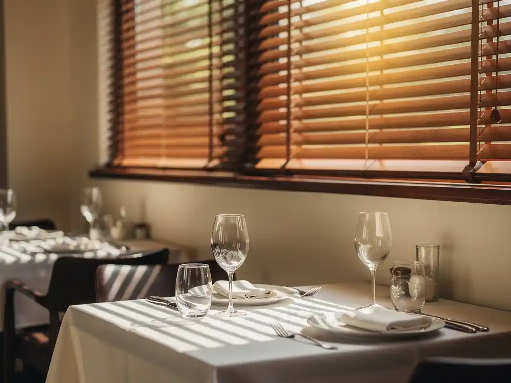 Houten veneziaanse jaloezieen filteren warm middaglicht in een stijlvol restaurant met linnen tafels en wijnglazen.